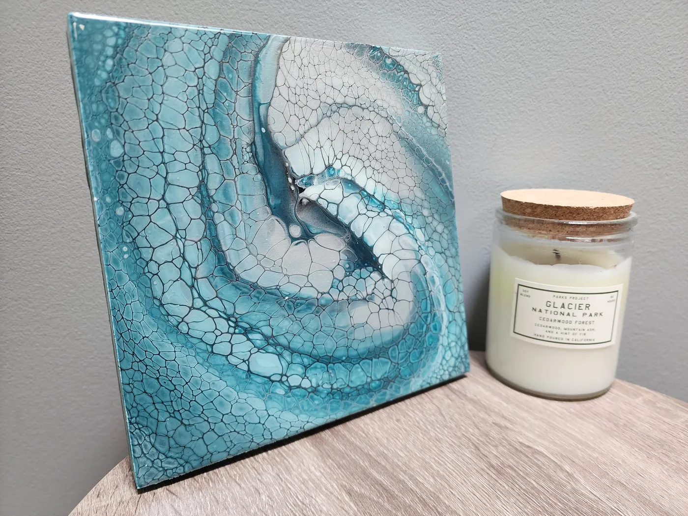 A small, square fluid art piece with swirling turquoise, grays, and whites on an end table next to a candle