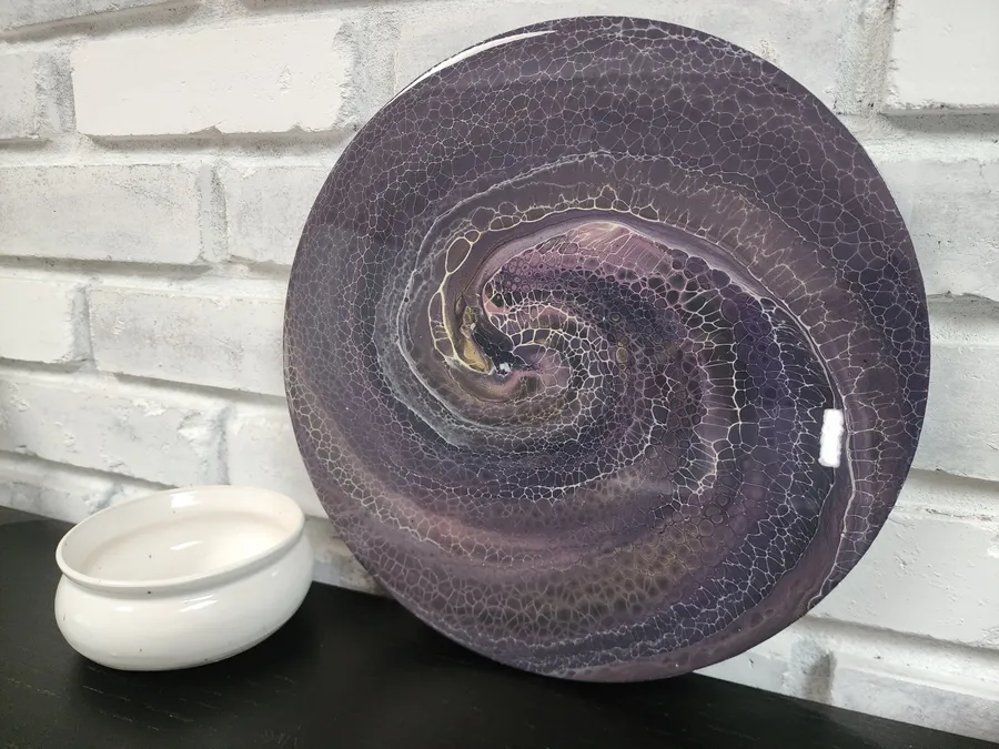 A round fluid art piece on a black shelf with a candle sitting beside it. The art piece is a swirling purple, gray, and metallic gold.