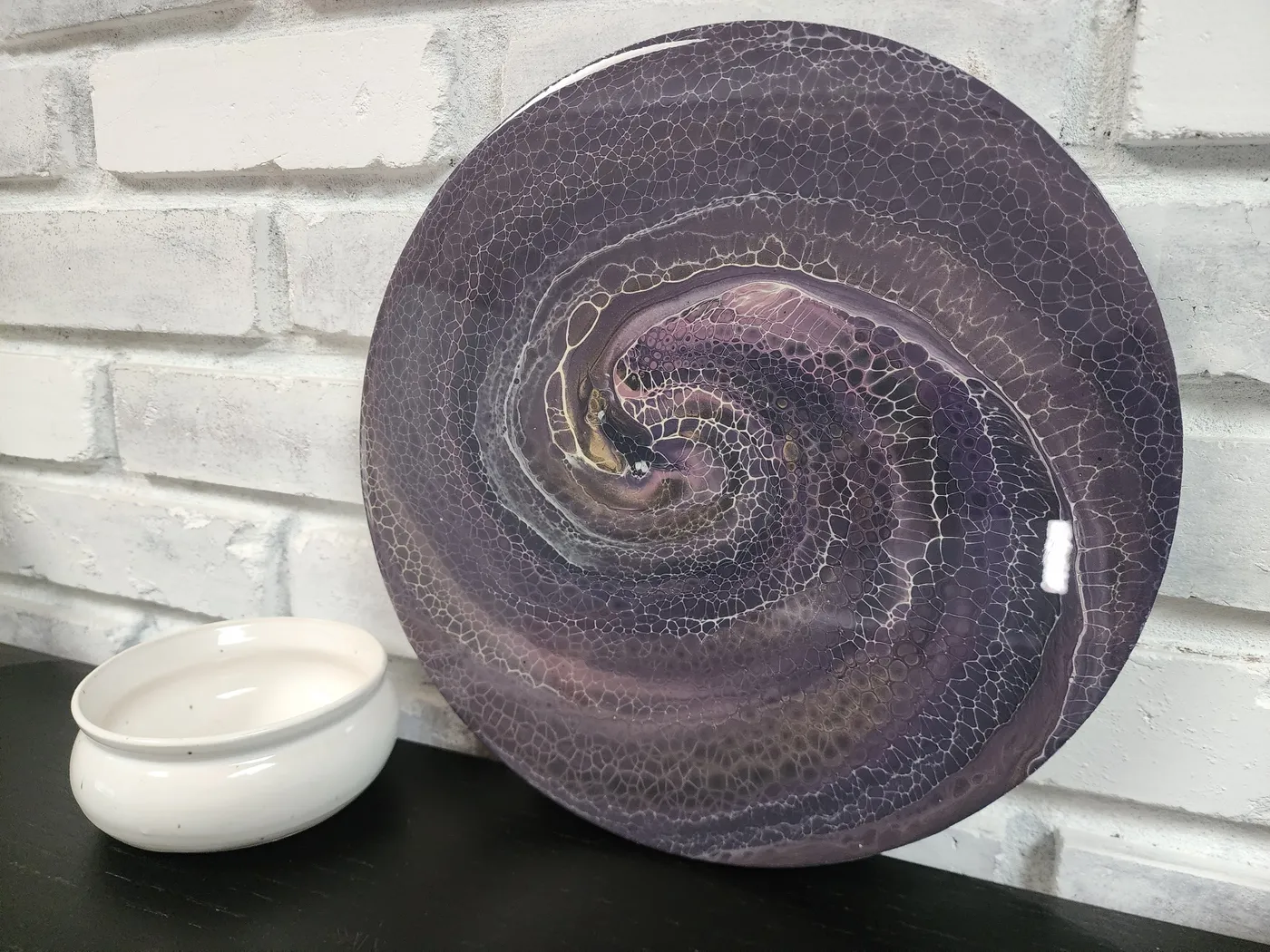 A round fluid art piece on a black shelf with a candle sitting beside it. The art piece is a swirling purple, gray, and metallic gold.