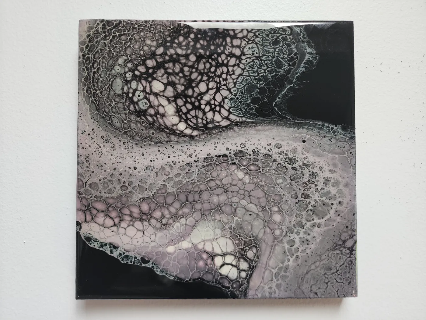 A small, square fluid art piece on a neutral wall. The piece shows off a wave of pale lavender and gray fluid cells over a black base.