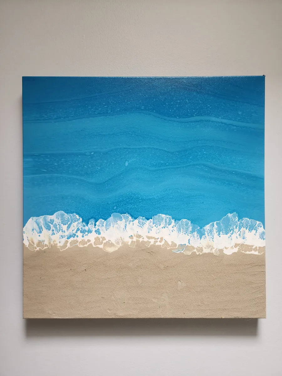 A square canvas fluid painting of a seascape from above. The piece has a striking blue ocean with foaming waves where it meets the beach made of real sand on canvas.