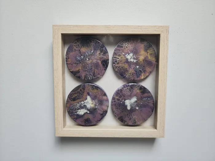 A wood-framed art piece on a neutral wall. The frame is a light wood with a glass pane. Four wooden round fluid art pieces are mounted evenly in a square pattern in the frame behind the glass. Each round showcases Purple, gold, and white fluid art cell designs over a black base.