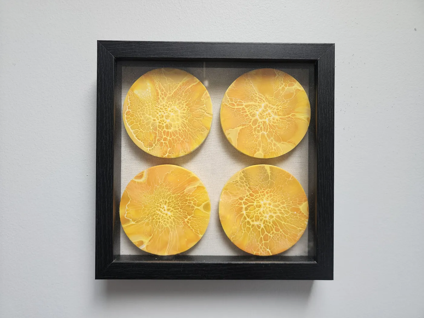 A wood-framed art piece on a neutral wall. The frame is painted black wood with a glass pane. Four wooden round fluid art pieces are mounted evenly in a square pattern in the frame behind the glass. Each round showcases yellow, orange, and golden fluid art cell designs over a white base.