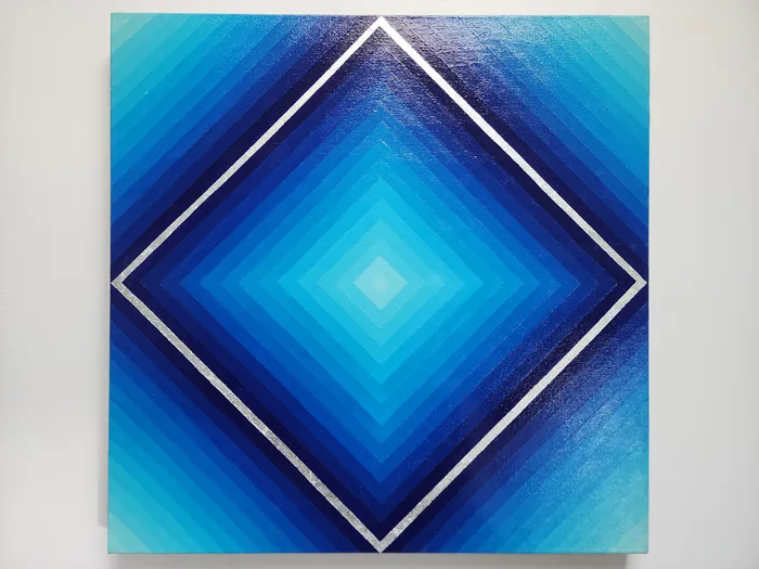 An acrylic canvas painting on a neutral wall. The piece showcases a clean-line blue gradient in a diamond pattern. This gradient starts with a square of silver leaf, then fades in each direction to a light blue in the middle.