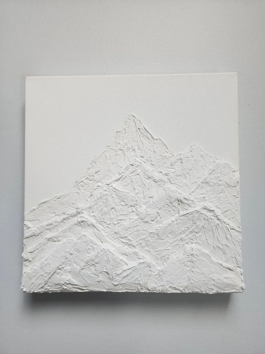An unpainted, white canvas piece on a neutral wall. The piece is a textured art piece made of plaster depicting layered mountain peaks.