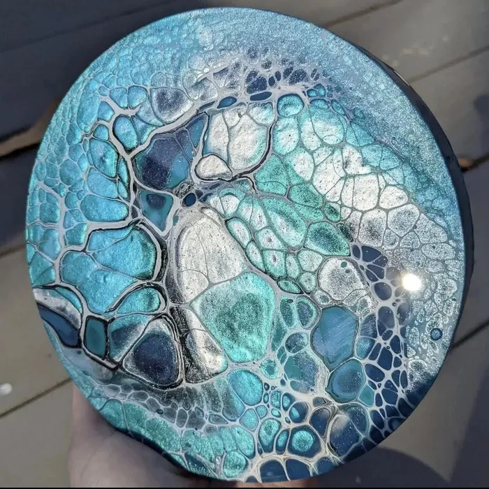 Round canvas with a teal and silver acrylic pour, held against weathered decking; large cells edged in dark blue spread across the surface.