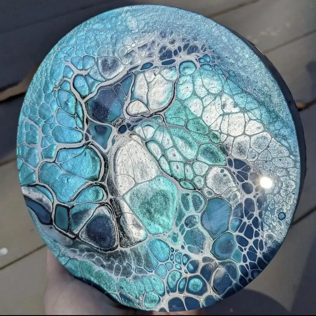 Round canvas with a teal and silver acrylic pour, held against weathered decking; large cells edged in dark blue spread across the surface.