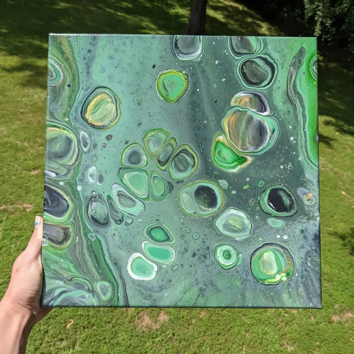 Square canvas held up over grass, acrylic poured in deep green with pale cells ringed in gold and white.