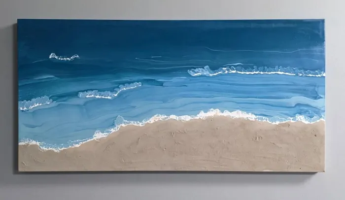 Wide canvas on a grey wall showing an aerial shoreline: deep blue water breaking into white surf against a broad band of real sand.