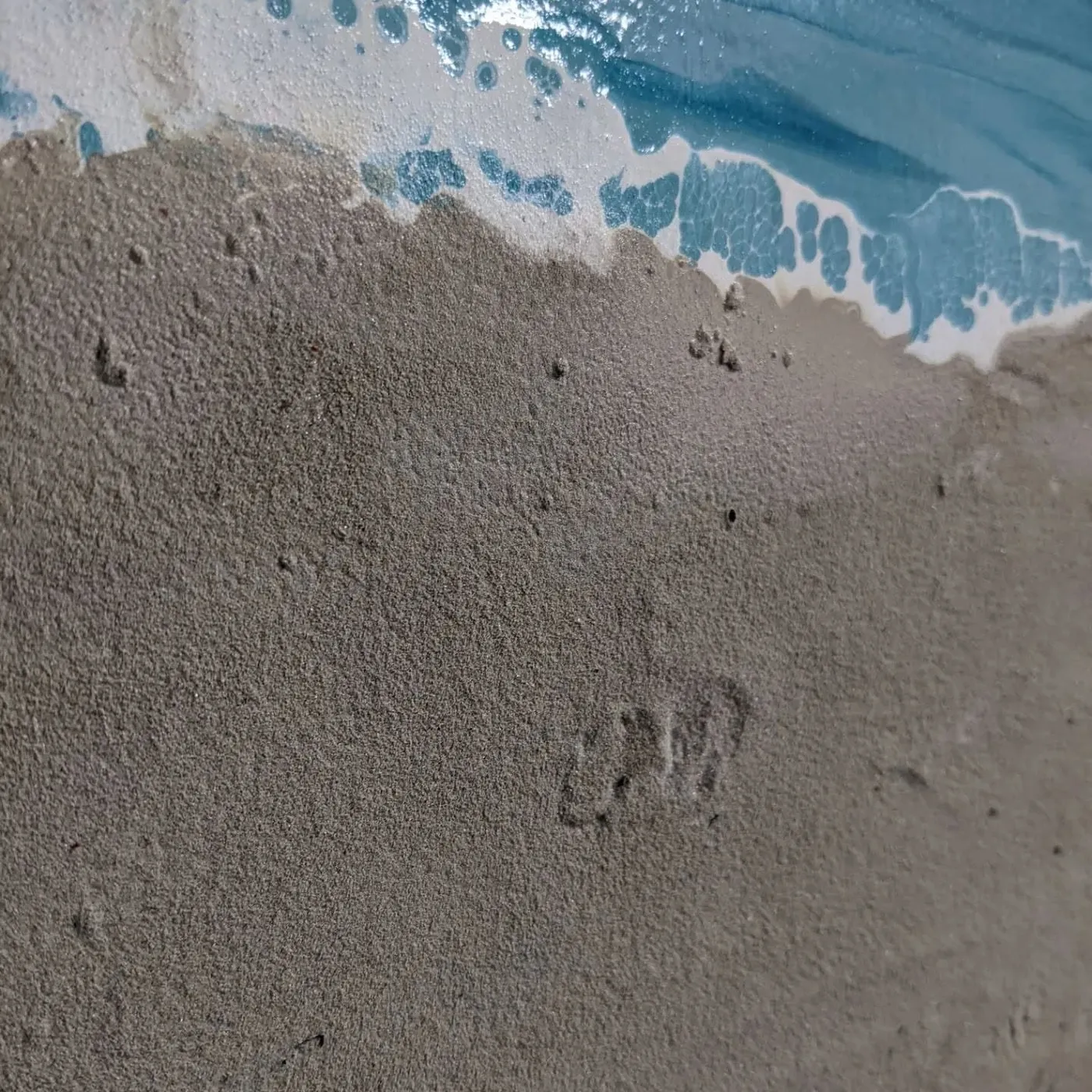 Close view where sand meets the water line, the grain of the real sand visible against glossy blue.