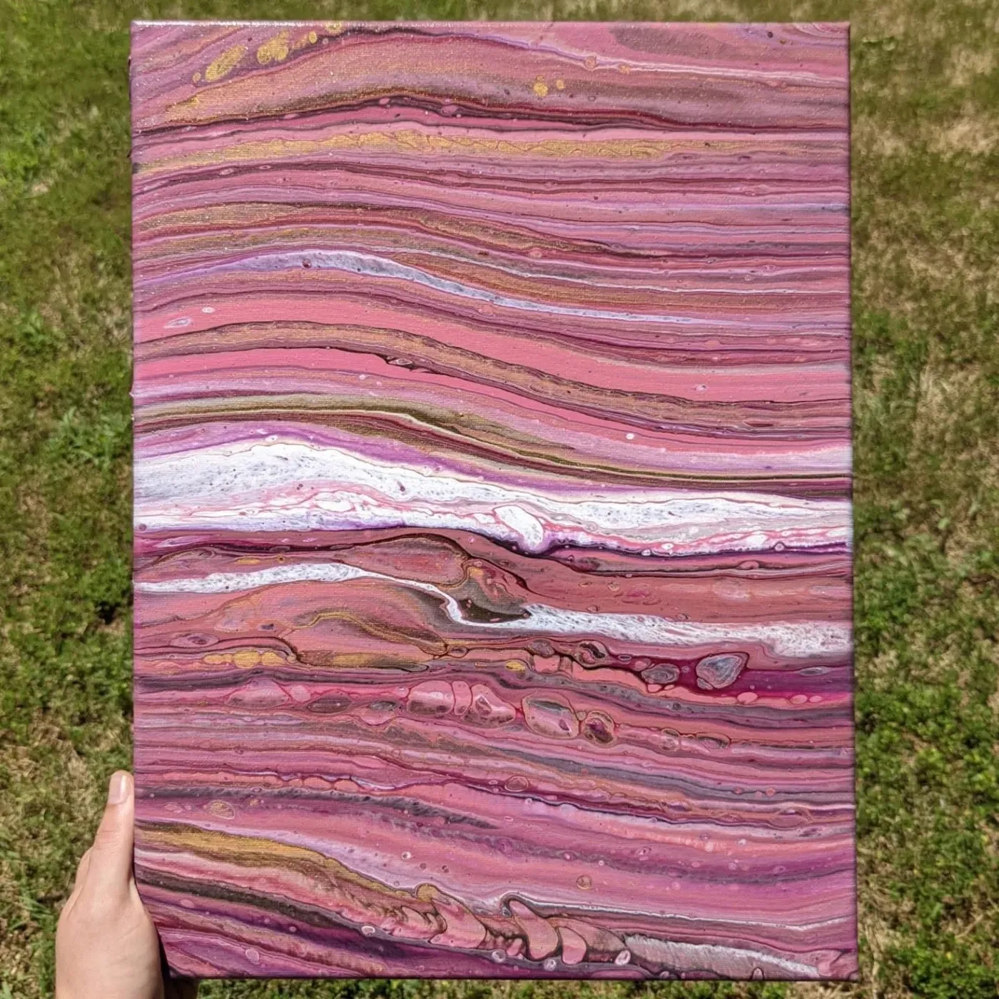 Portrait canvas held over grass, acrylic poured in rippling horizontal bands of pink, white and gold.